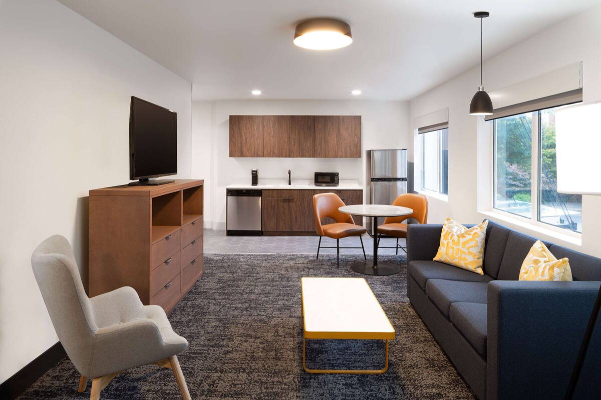 A modern living room with a flat-screen TV, a sofa with patterned cushions, a coffee table, and a dining area with a round table and chairs.