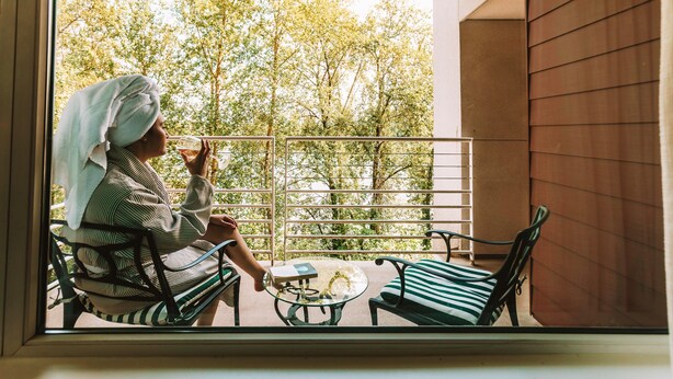 A balcony with a person sitting on a chair, holding a glass. There is a table with a book and a decorative item. The balcony has a metal railing and a view of trees. - River's Edge Hotel Portland, Tapestry Collection by Hilton (Portland)