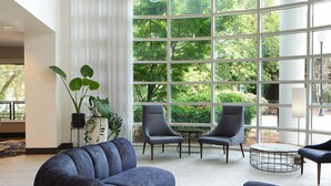 A modern living room with a sectional sofa, two armchairs, and a coffee table. Large windows offer a view of greenery outside. - River's Edge Hotel Portland, Tapestry Collection by Hilton (Portland)