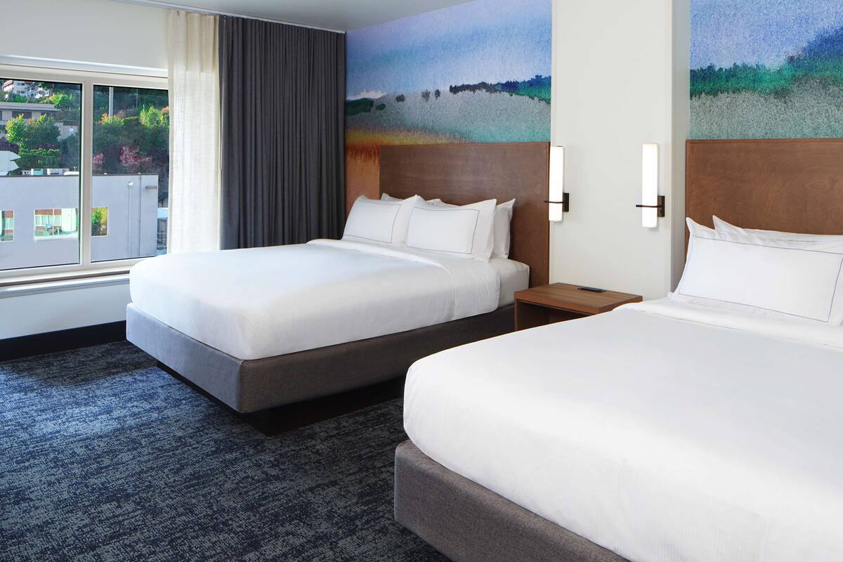 A hotel room with two beds, a large window with curtains, and a painting on the wall.