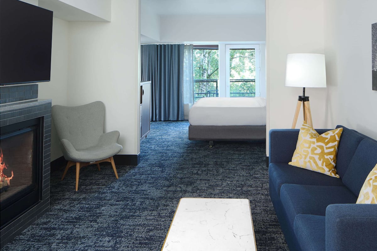 A hotel room with a fireplace, a sofa, a chair, a bed, and a large window with curtains.