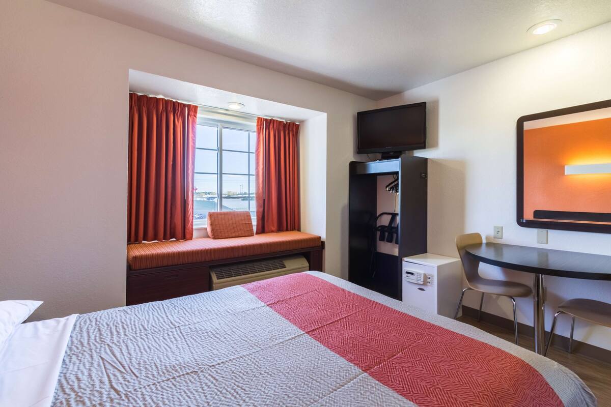 Habitación de hotel con cama, escritorio, silla, televisor y ventana con cortinas.