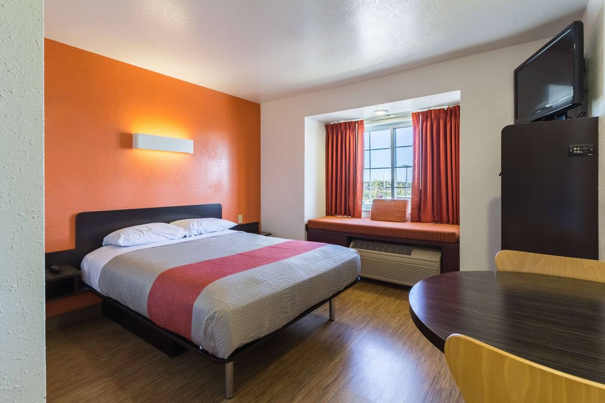 Habitación de hotel con cama, escritorio, silla, televisor y ventana con cortinas.