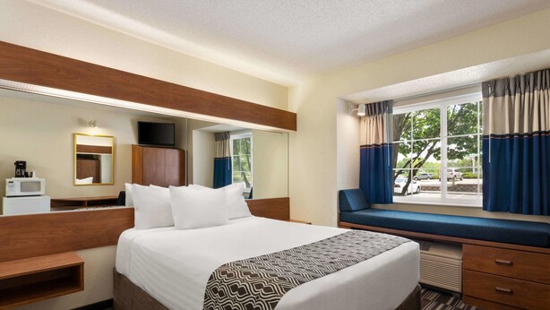Habitación de hotel con una cama grande, una mesita de noche, un espejo, un televisor, una ventana con cortinas y vistas al exterior.