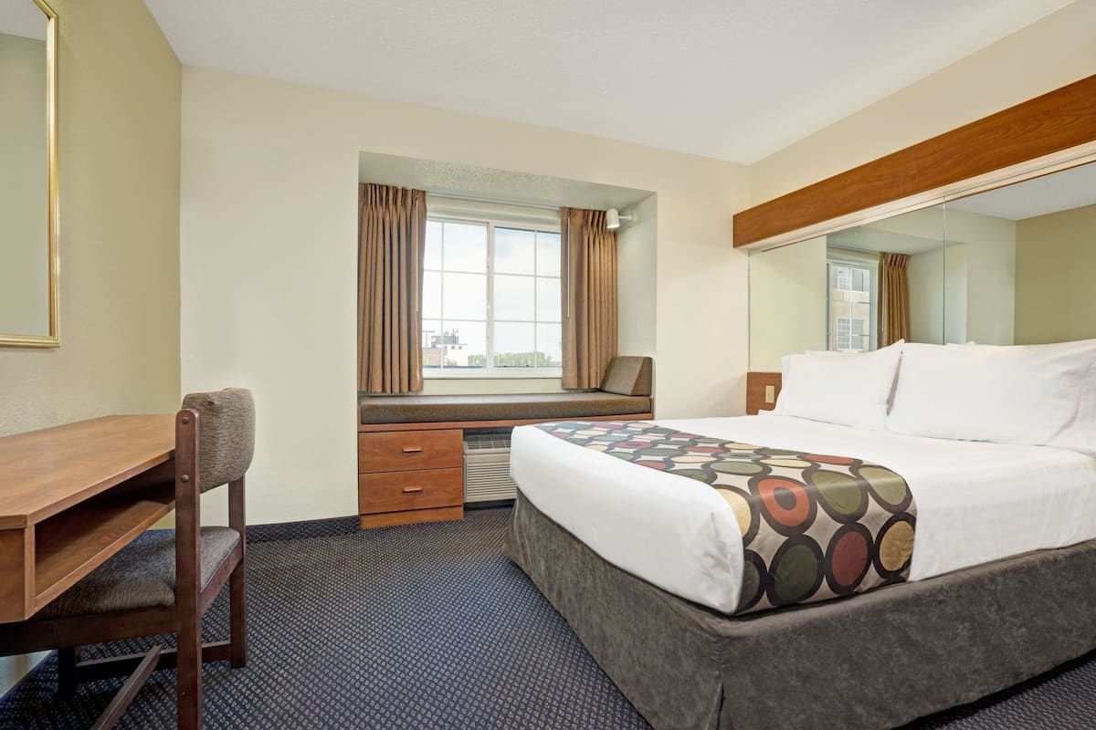 A hotel room with a bed, a desk, a chair, a window with curtains, and a mirror.