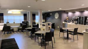 Un comedor de hotel moderno con mesas y sillas, una zona de cocina con electrodomésticos y un gran reloj en la pared.