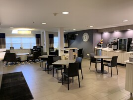 Un comedor de hotel moderno con mesas y sillas, una zona de cocina con electrodomésticos y un gran reloj en la pared.