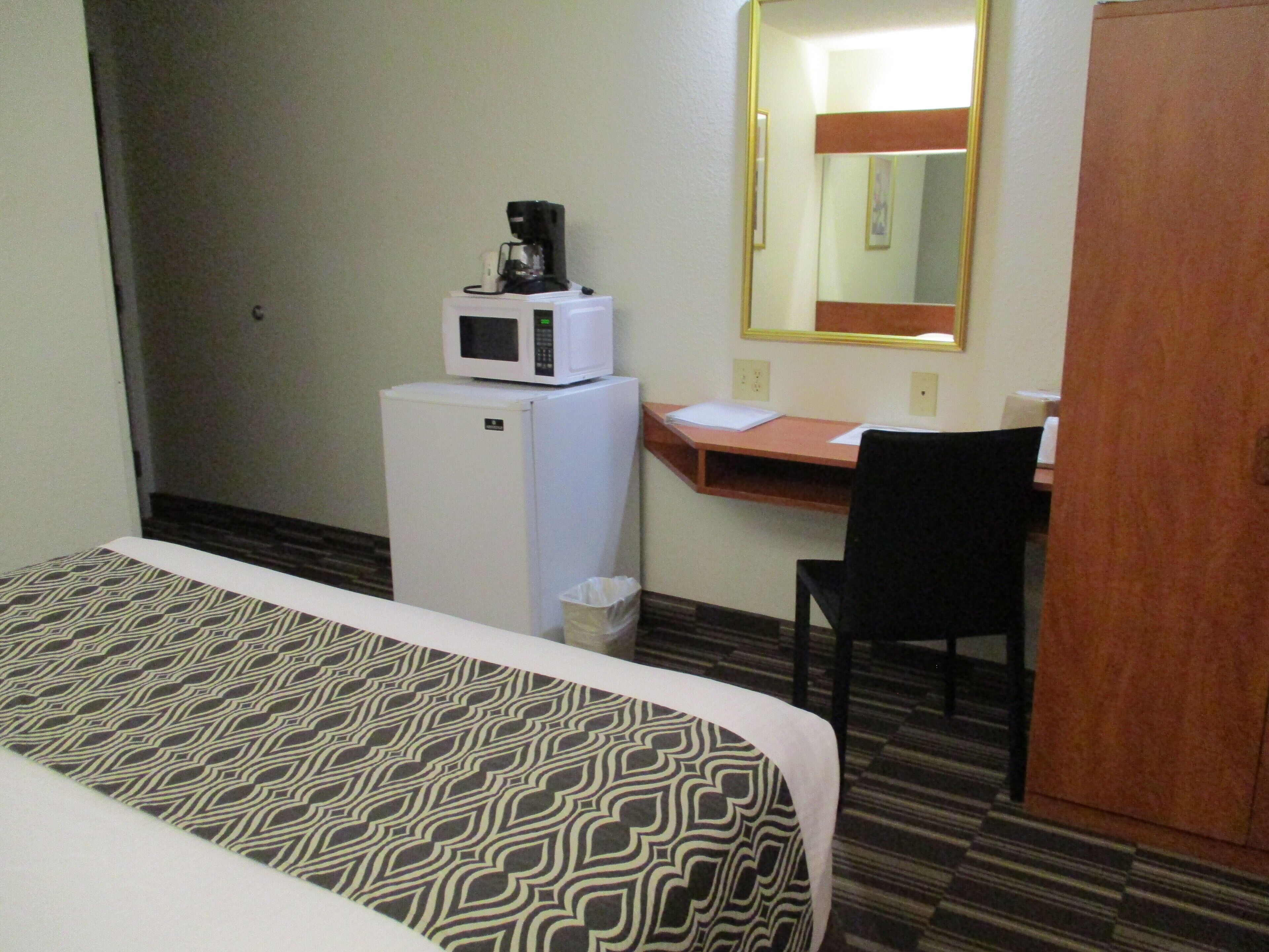 Quarto de hotel com uma cama, uma escrivaninha com cadeira, micro-ondas sobre um mini-refrigerador e um espelho na parede.