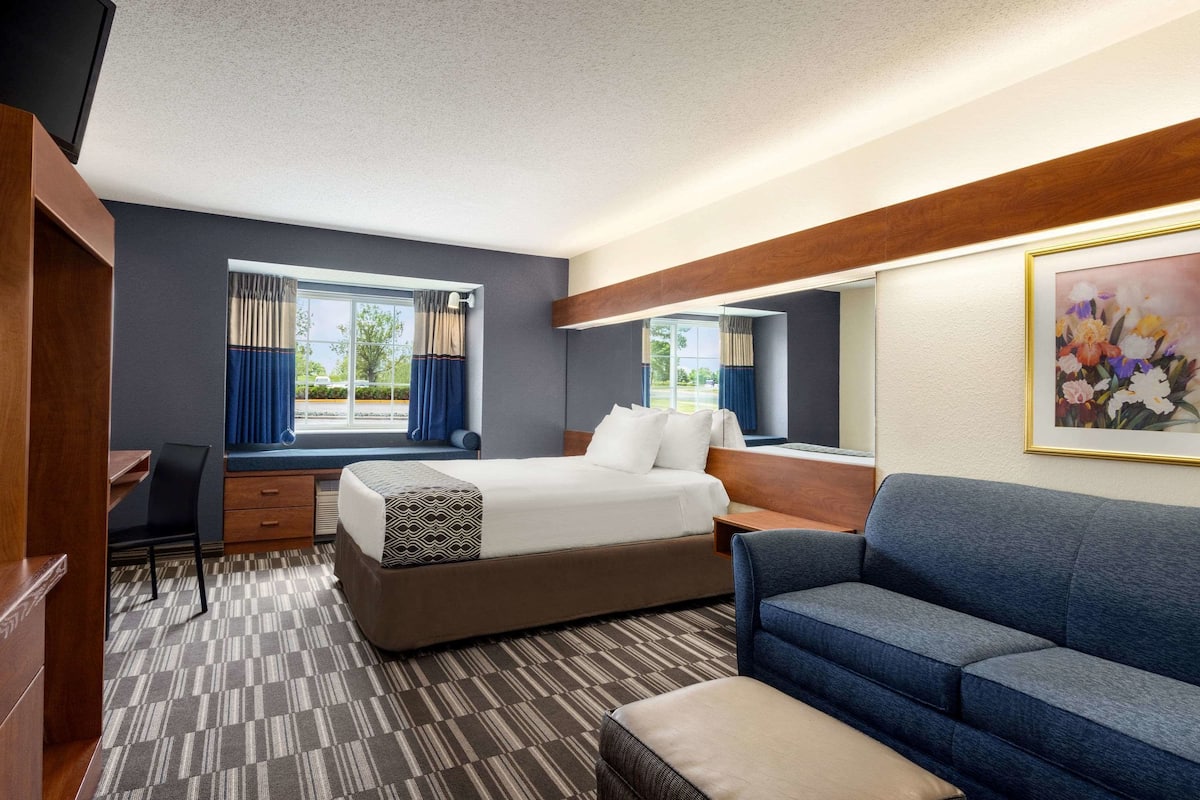 A hotel room with a bed, a desk, a chair, a sofa, and a large window with curtains.