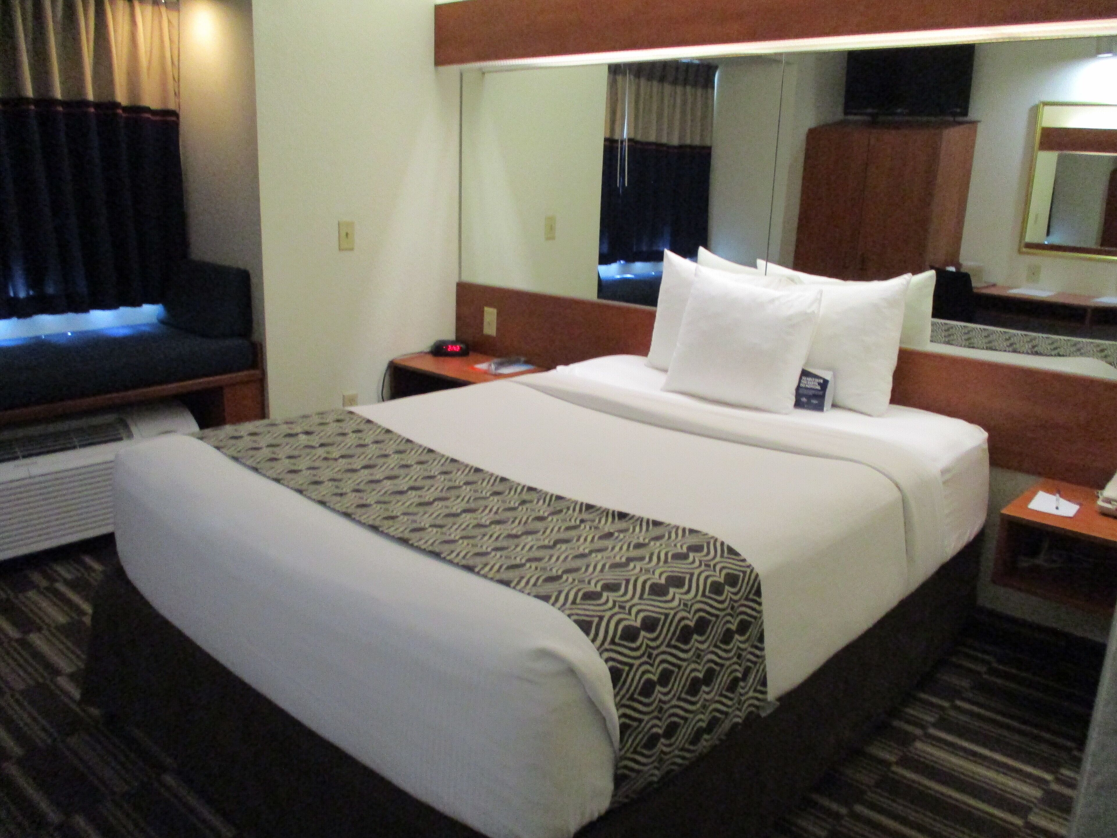 A hotel room with a bed, a bedside table, a nightstand, a mirror, a TV, and a window with curtains.