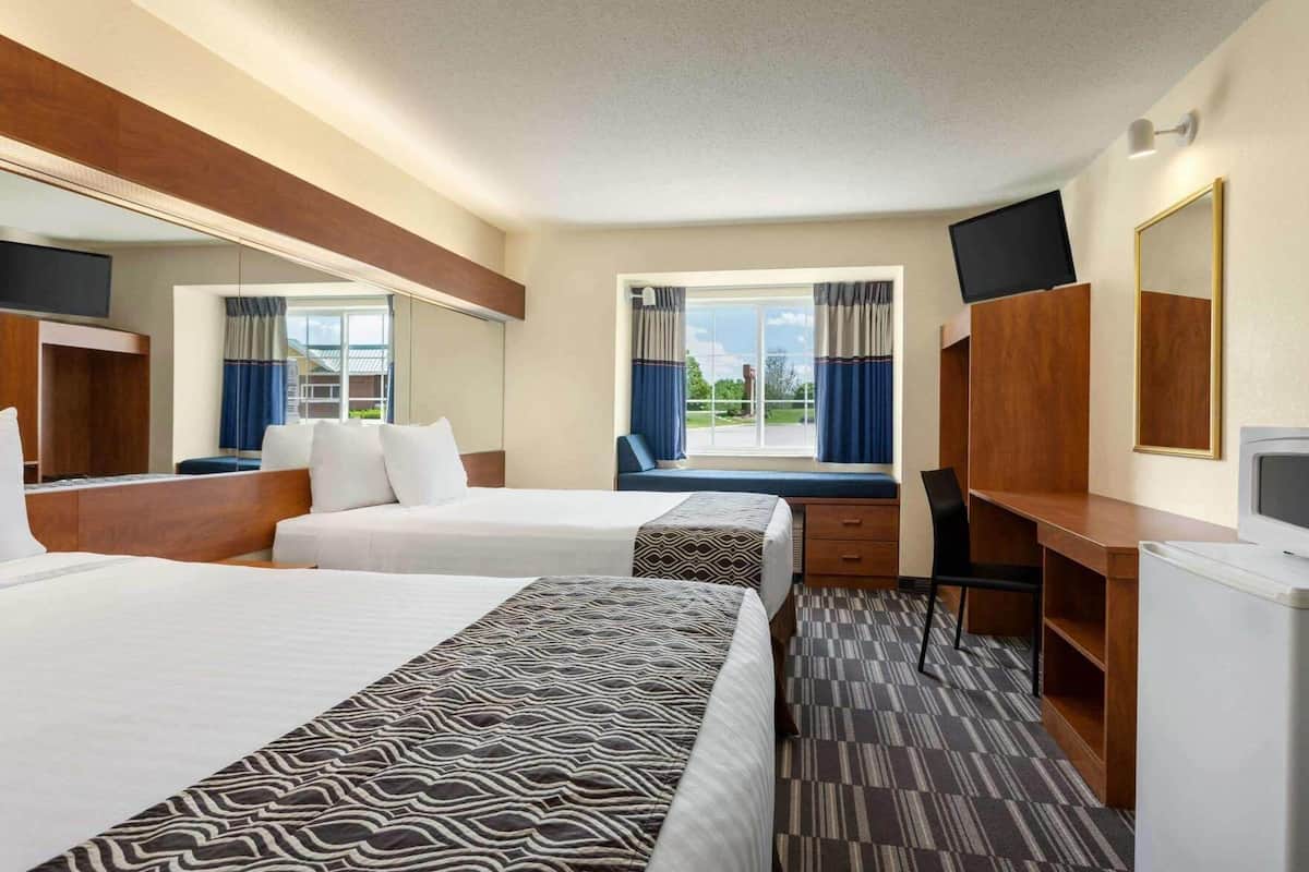A hotel room with two beds, a desk, a chair, a TV, a mirror, and a window with curtains.