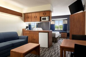 Habitación de hotel compacta con kitchenette, sofá, mesa de comedor y refrigerador pequeño.