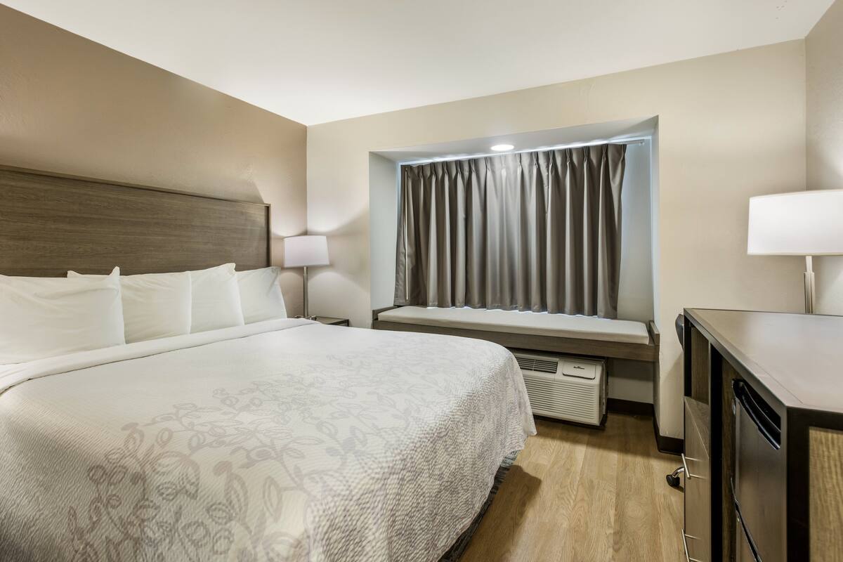 A hotel room with a large bed, a bedside table with lamps, a window with curtains, and a built-in bench.