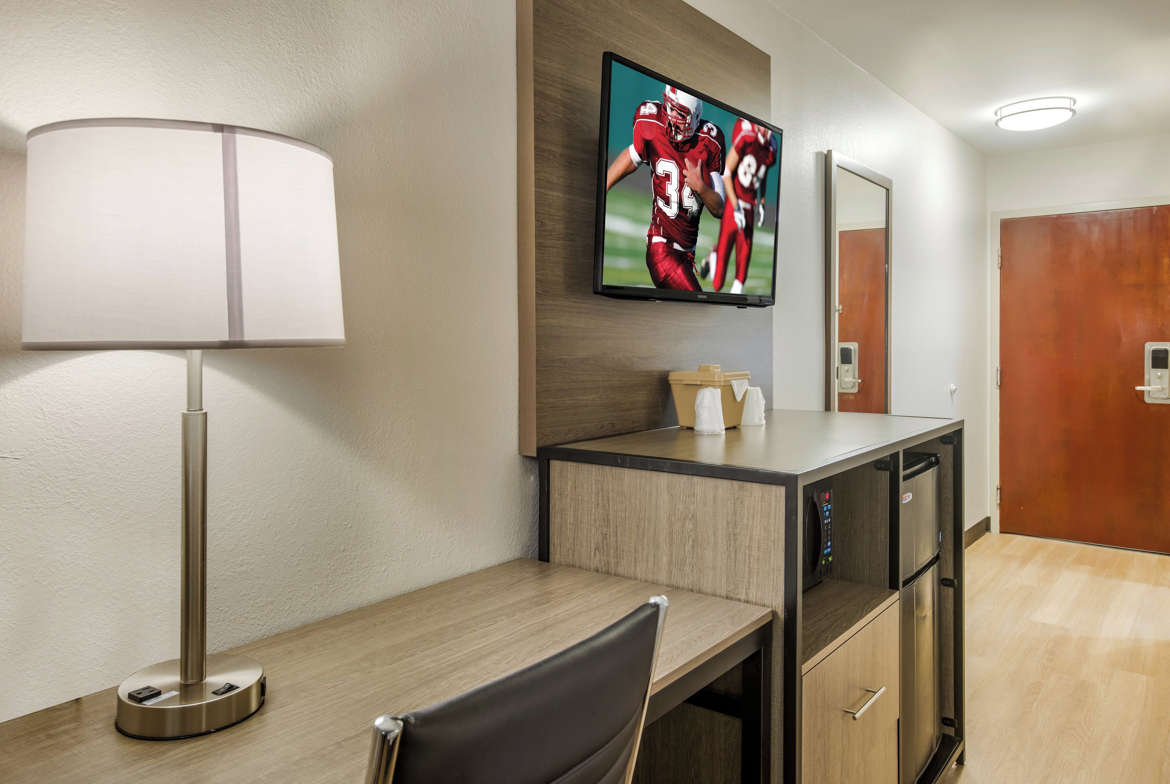 Habitación de hotel con una televisión de pantalla plana montada en la pared, un escritorio con una lámpara, una silla y un mueble de madera con microondas.