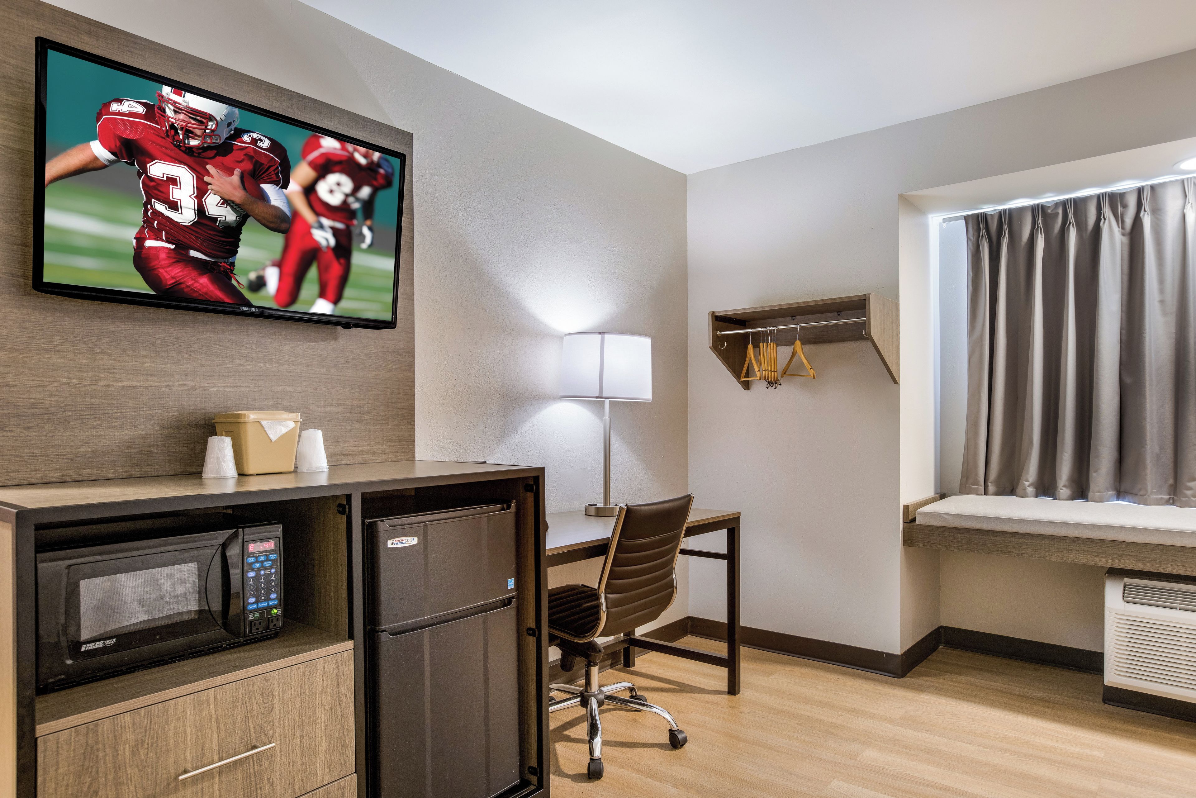 Quarto de hotel com televisão na parede, uma escrivaninha com cadeira, micro-ondas, frigorífico, janela com cortinas e prateleira com cabides.