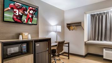 Quarto de hotel com televisão na parede, uma escrivaninha com cadeira, micro-ondas, frigorífico, janela com cortinas e prateleira com cabides.