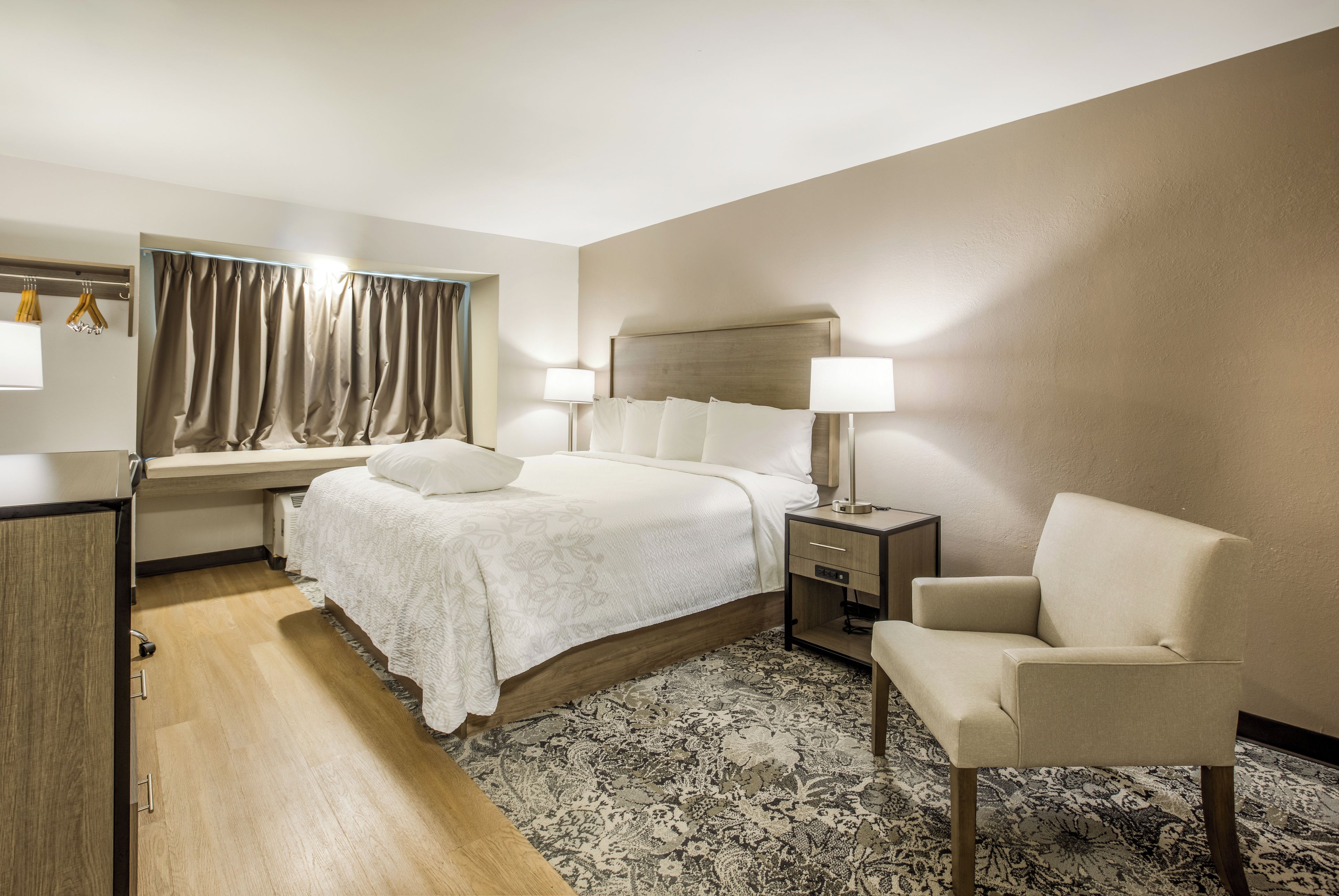 Una habitación de hotel con una cama, mesitas de noche, una silla, un escritorio y una ventana con cortinas.