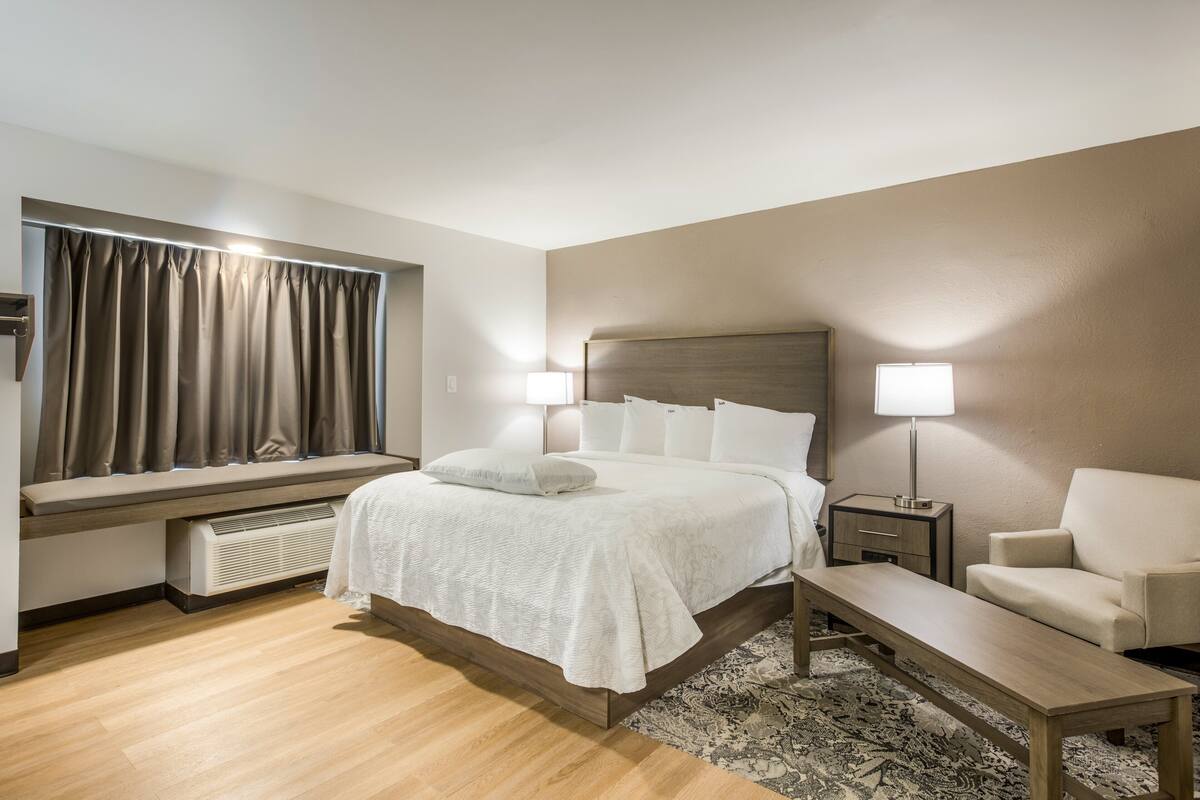 A hotel room with a bed, bedside tables, a bench, a sofa, a window with curtains, and a radiator.