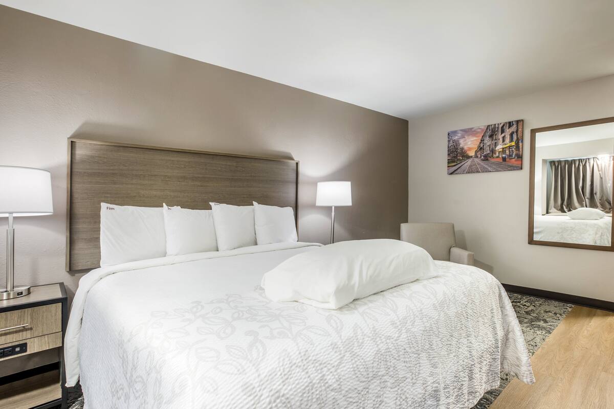 A hotel room with a large bed, a nightstand, a lamp, a painting, and a mirror.
