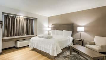 A hotel room with a bed, bedside lamps, a chair, a small table, a window with curtains, and a radiator.