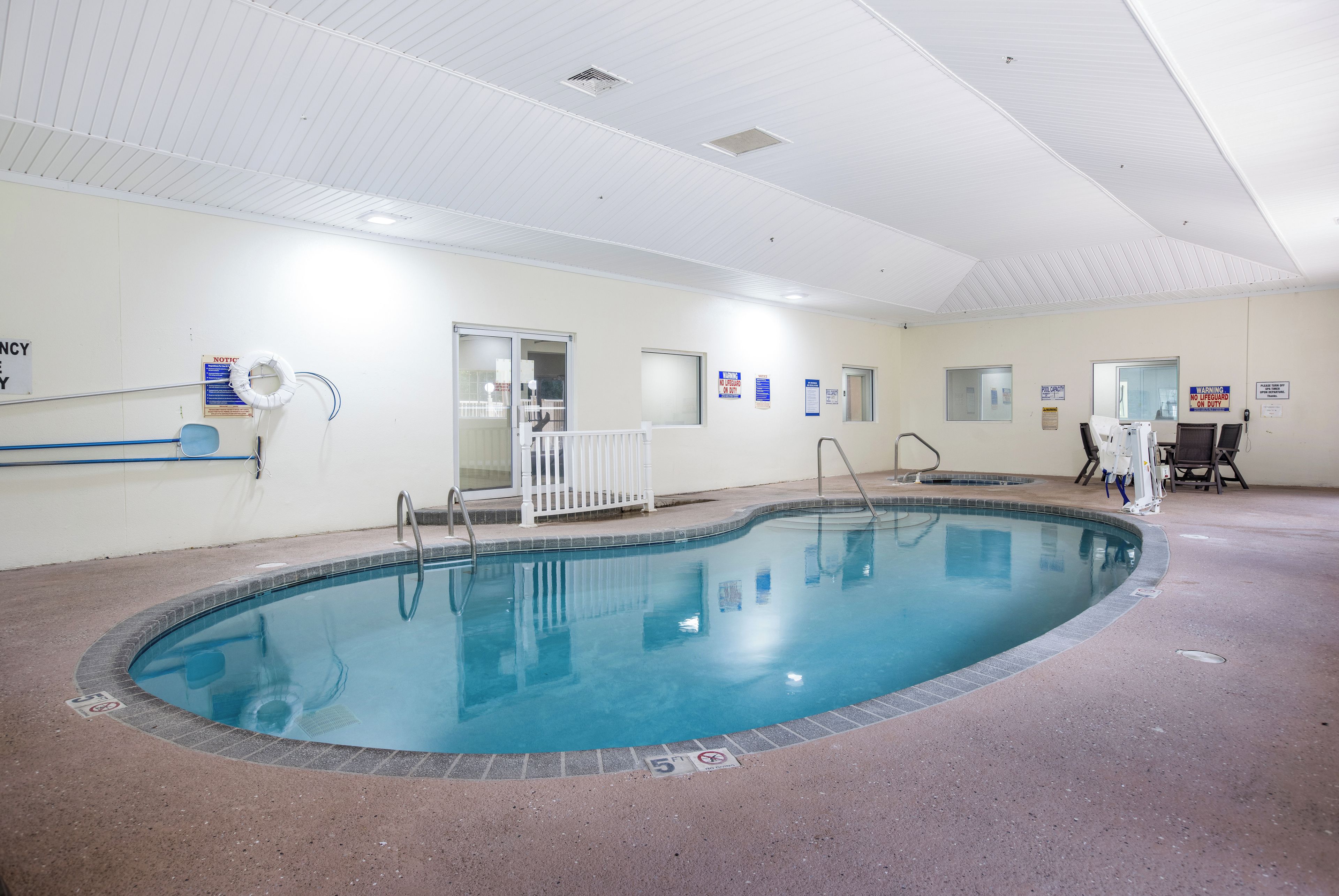 An indoor swimming pool with a clear water surface, surrounded by a tiled floor and walls. There are signs on the walls, a white railing around the pool, and a door leading to another room.