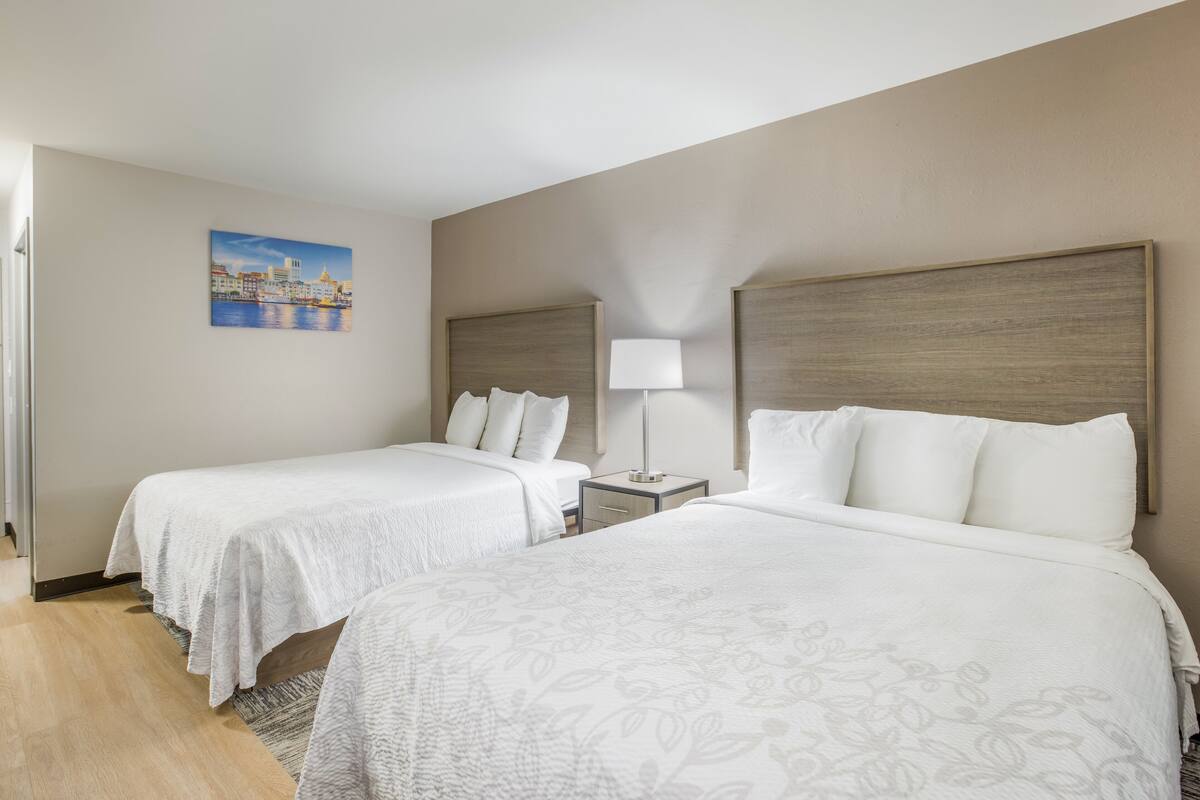 A hotel room with two beds, a nightstand with a lamp, a painting on the wall, and a wooden floor.