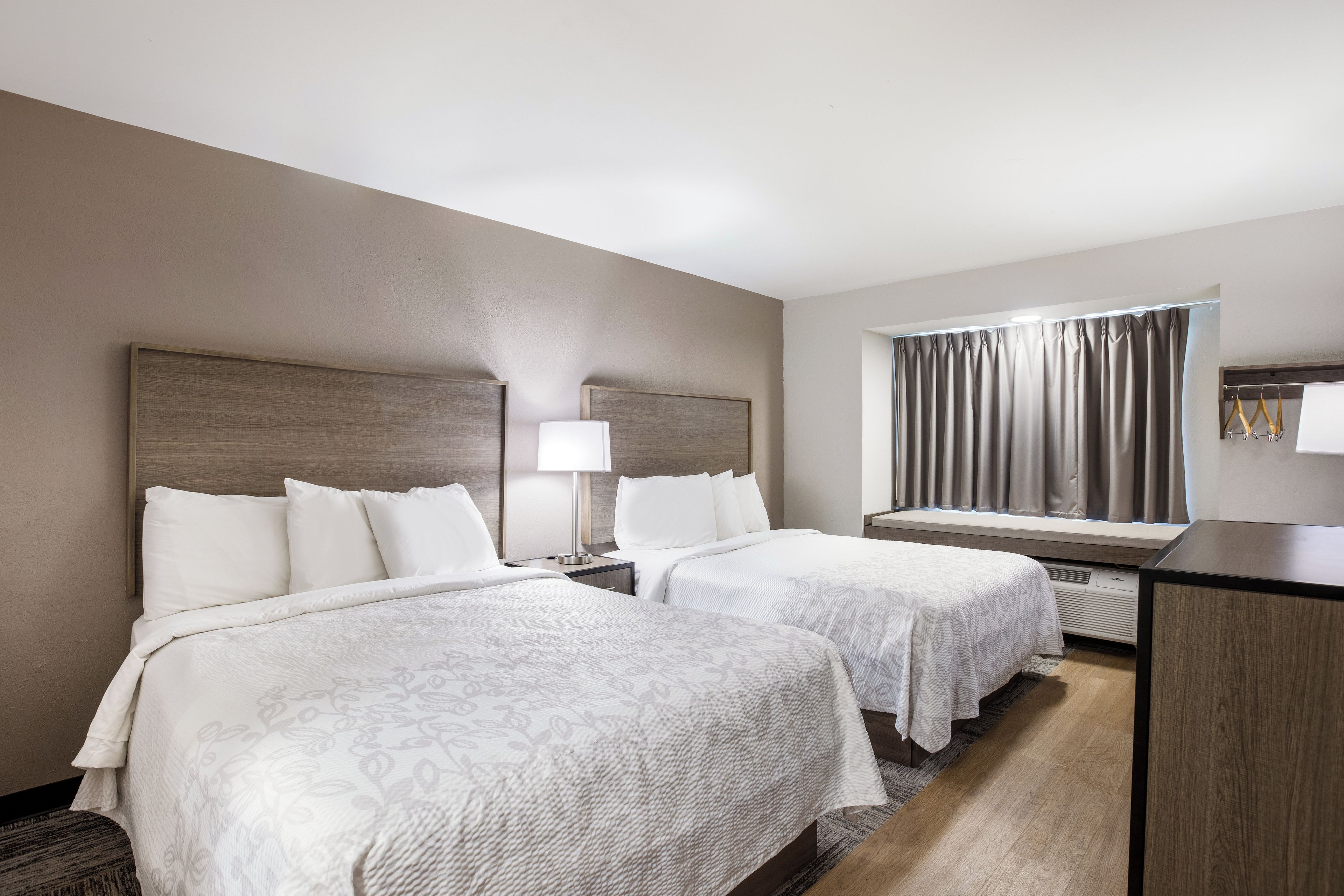 A hotel room with two beds, a desk, a window with curtains, and a wall-mounted heater.