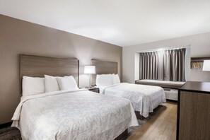 A hotel room with two beds, a desk, a window with curtains, and a wall-mounted heater.