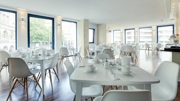 Une salle à manger spacieuse, avec des tables et des chaises blanches, de grandes fenêtres et une vue sur les bâtiments environnants.