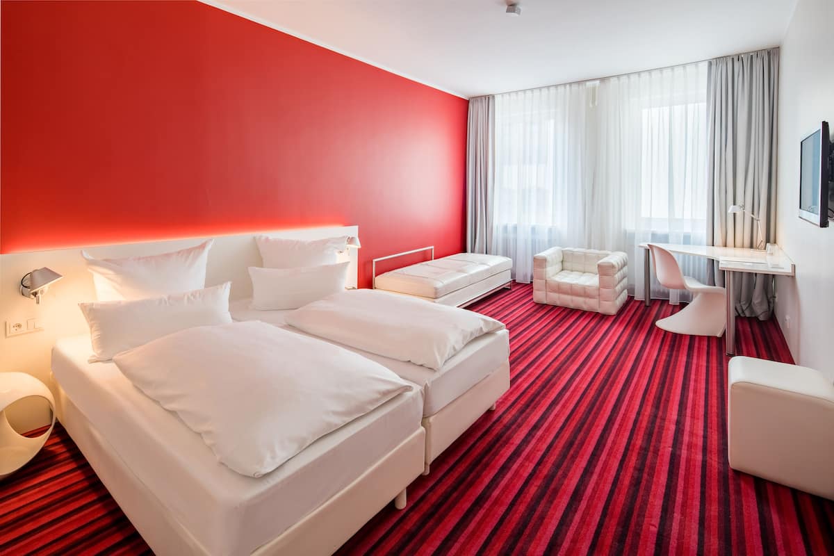A hotel room with a red carpet, a bed, a desk, and a chair.