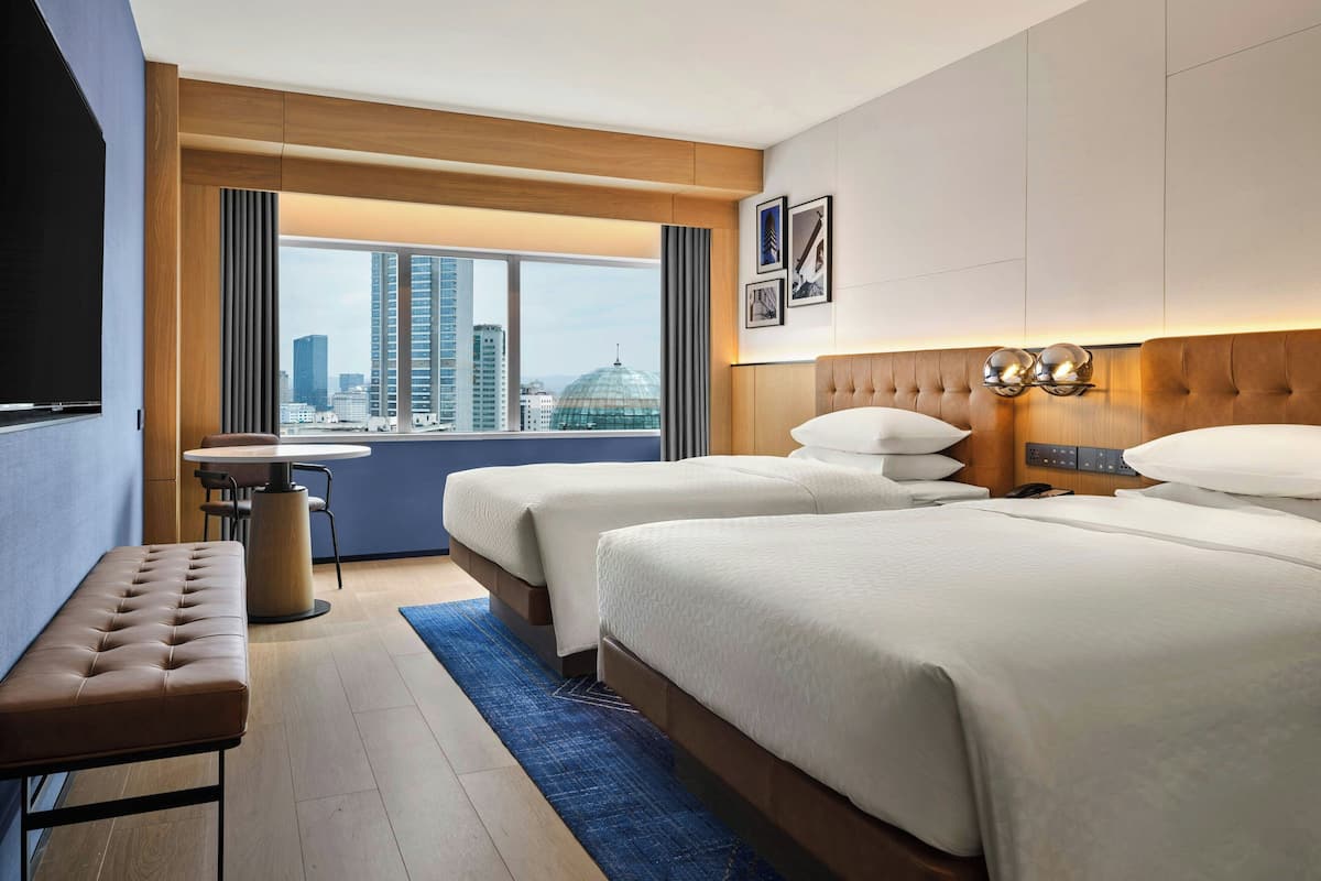 Une chambre d’hôtel avec deux lits, une banquette, une petite table et une vue sur le paysage urbain.