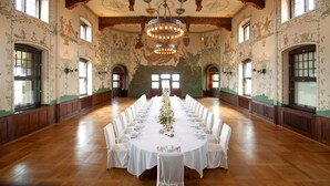 Ein langer Festsaaltisch ist für eine formelle Veranstaltung gedeckt, umgeben von weißen Stühlen, in einem Raum mit aufwendigen Wandmalereien und großen Fenstern.