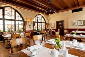 Un restaurante con mesas y sillas de madera, amplios ventanales en arco y un candelabro.
