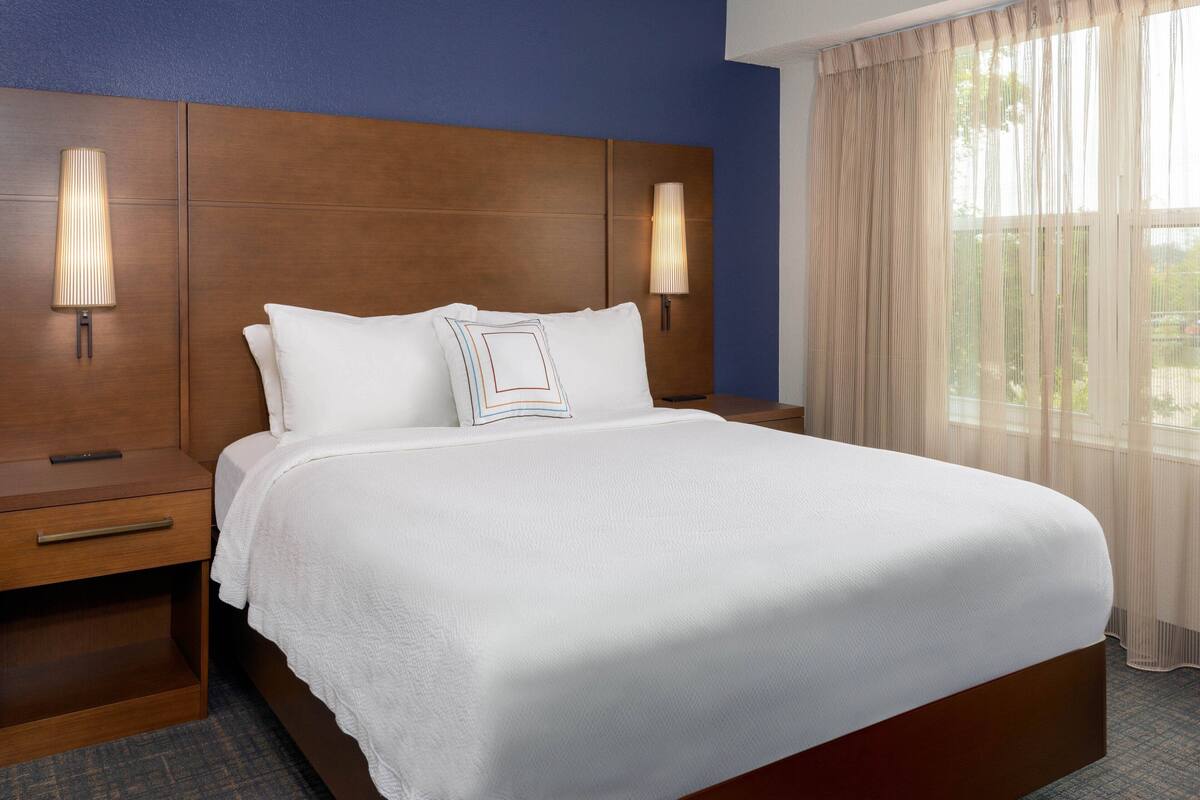 Een hotelkamer met een groot bed, twee bedlampen, een houten hoofdbord en een raam met dunne gordijnen.