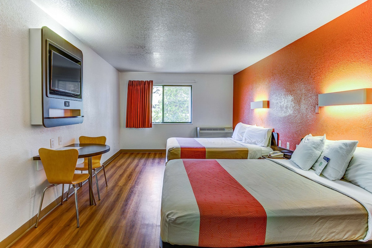 A hotel room with two beds, a dining table with chairs, a TV mounted on the wall, and a window with curtains.