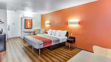 A modern hotel room with a wooden floor, a bed with white bedding, a desk with a chair, and a small table with a lamp.