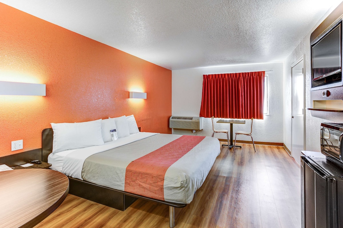 A hotel room with a bed, a desk, a chair, a TV, and a window with red curtains.