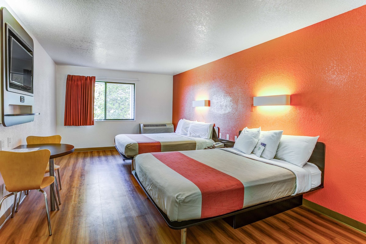 A hotel room with two beds, a dining table with chairs, a TV, and a window with curtains.
