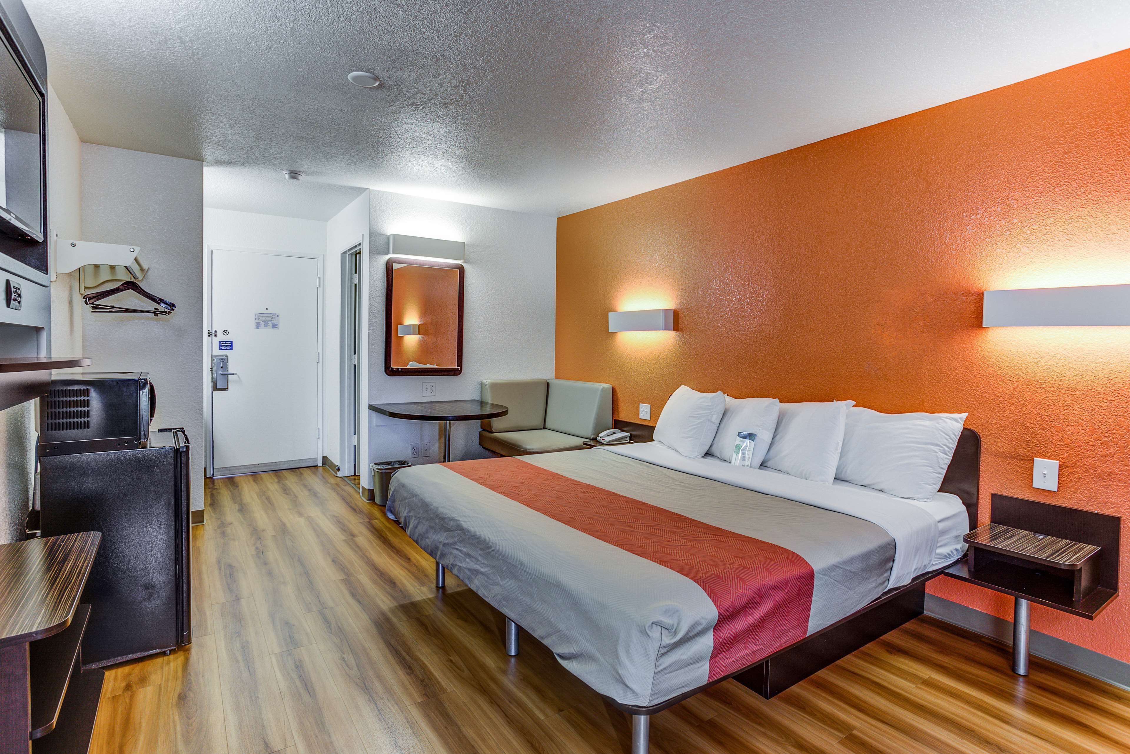 Habitación de hotel con cama, escritorio, silla, una mesita, espejo y lámpara en la pared.