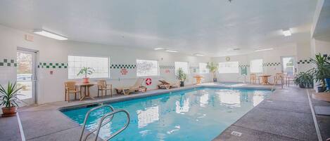 An indoor swimming pool with clear blue water, surrounded by lounge chairs and tables, with potted plants and a lifeguard sign on the wall.