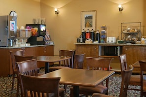 Un comedor de hotel con mesas y sillas de madera, una máquina de café, un refrigerador y un mostrador de buffet con alimentos.