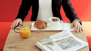 A person seated at a table with a croissant, a cup of coffee, and a glass of orange juice. A newspaper is also visible on the table.