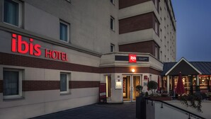 The exterior of an ibis hotel with a prominent red sign, a well-lit entrance, and outdoor seating area with umbrellas.