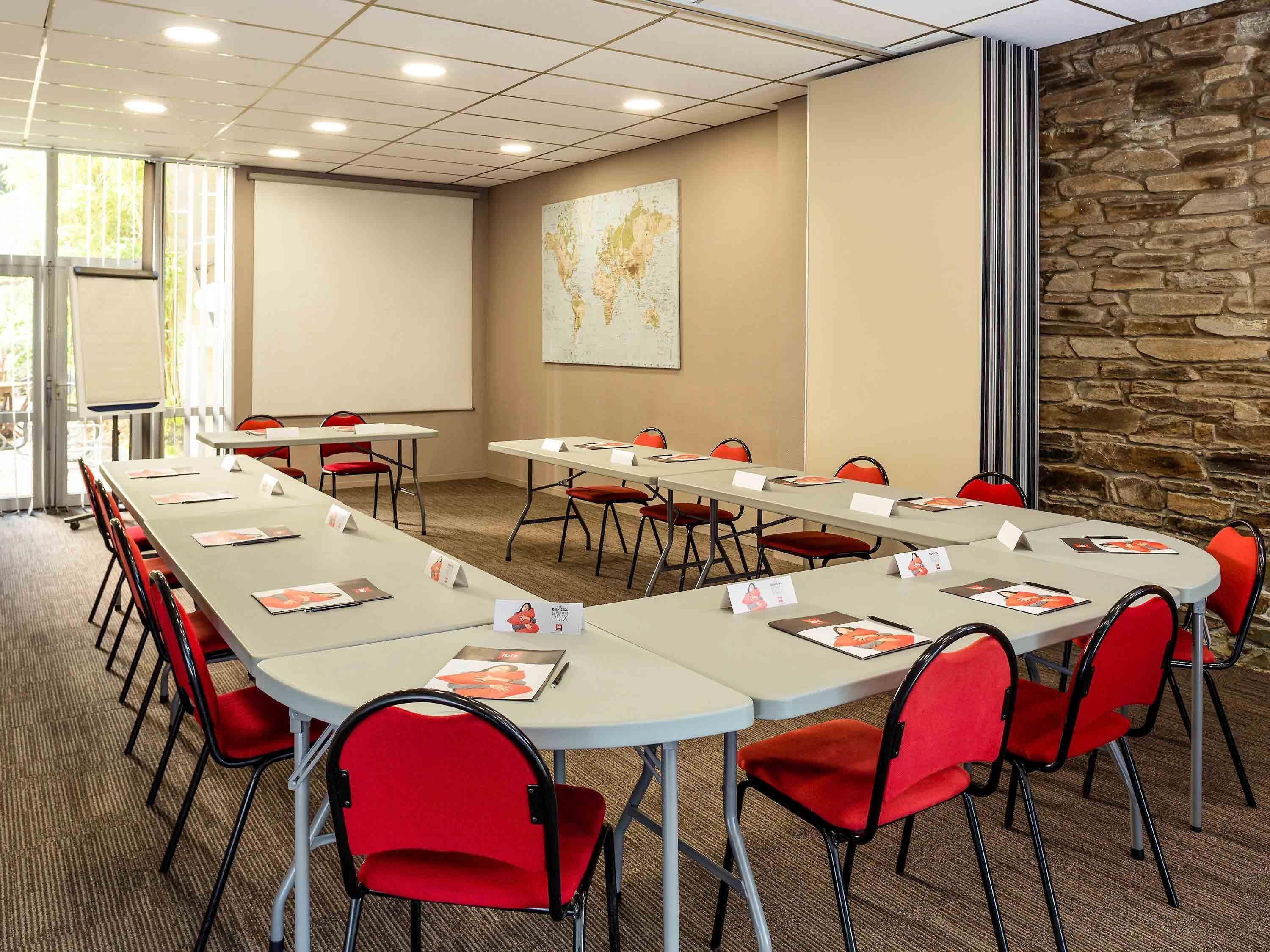 Une salle de conférence avec de longues tables, des chaises rouges, un tableau blanc et un mur en pierre.