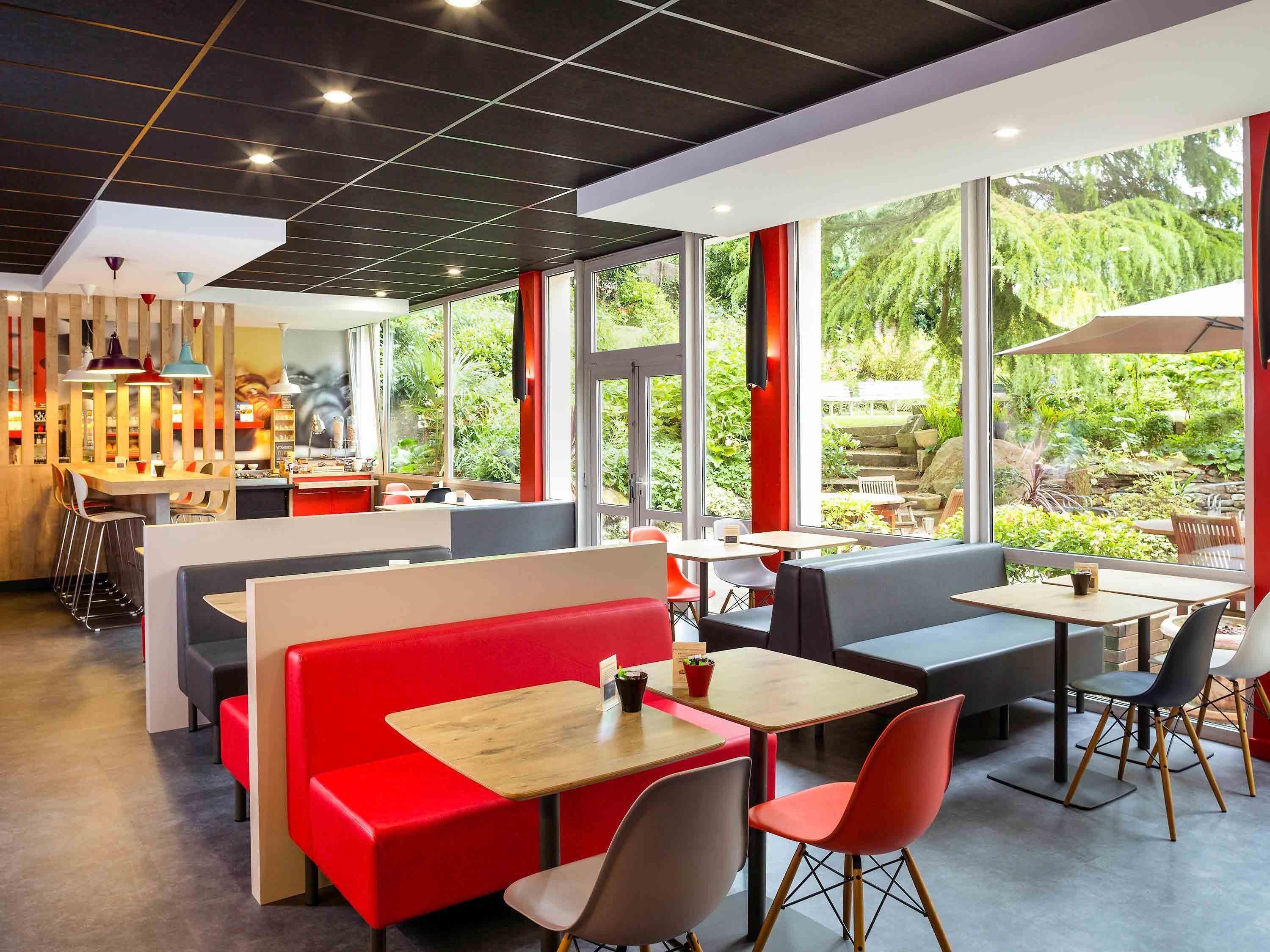 A modern cafe with red and grey seating, large windows offering a view of greenery, and a stylish ceiling design.