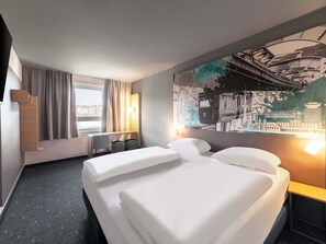 Une chambre d’hôtel avec deux lits, un bureau, une chaise, une grande fenêtre avec des rideaux et un tableau représentant un paysage urbain accroché au mur.