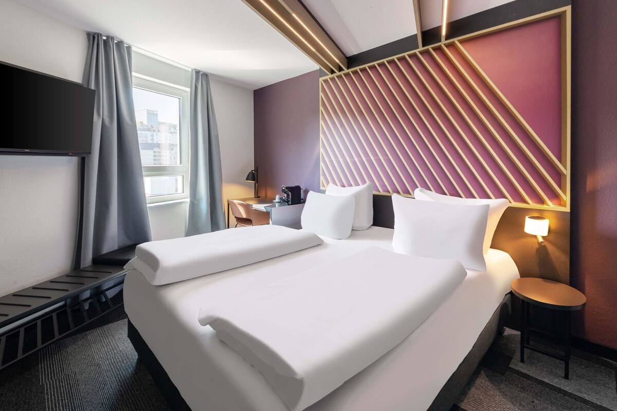 Ein Hotelzimmer mit Bett, Schreibtisch, Stuhl, Fernseher und einem Fenster mit Vorhängen.
