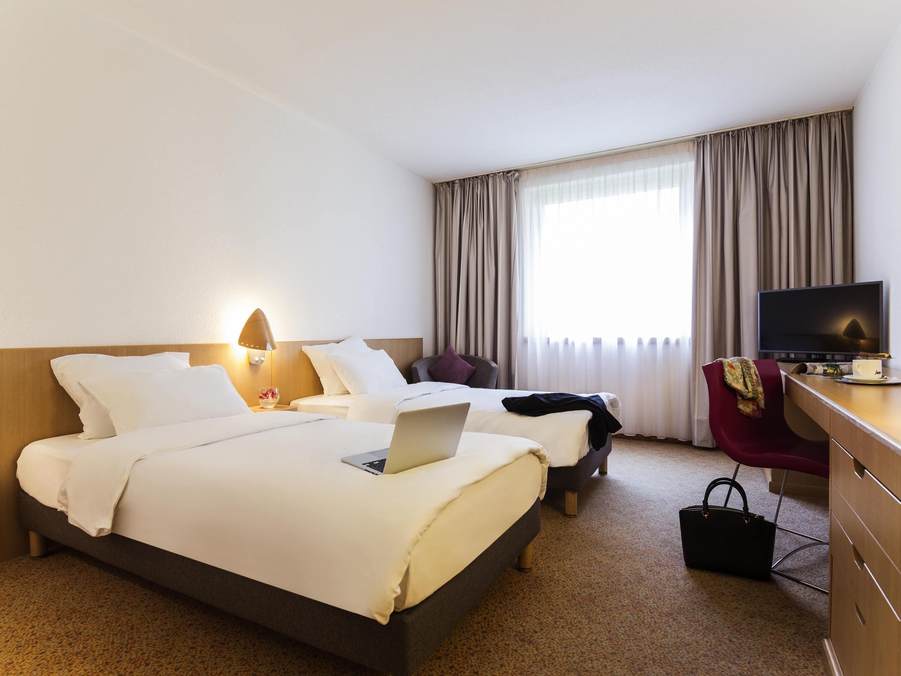 Ein Hotelzimmer mit zwei Betten, einem Schreibtisch mit Fernseher, einem Sessel und einer Schreibtischlampe.
