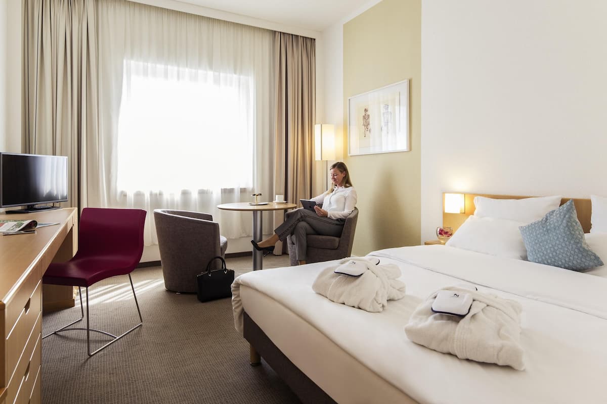 Une chambre d’hôtel comprenant un lit, un bureau avec une télévision, une chaise, une table ronde et une fenêtre avec des rideaux.