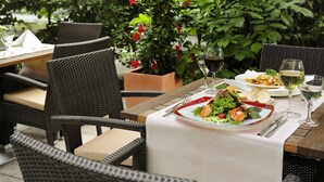 Un área de comedor al aire libre con sillas de mimbre, una mesa dispuesta con ensalada, copas de vino y una planta en maceta con flores rojas.